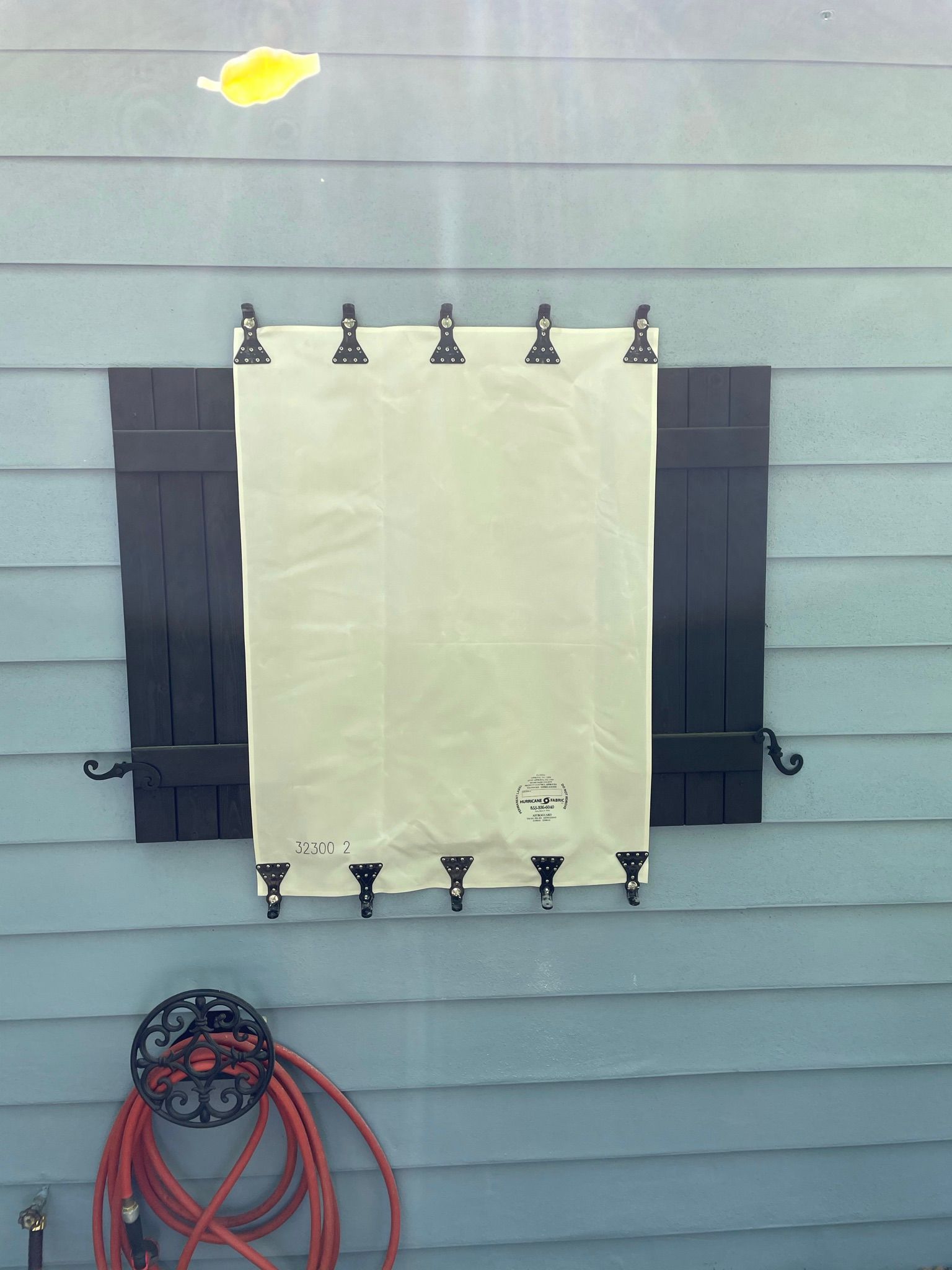 Blank beige tarp clipped to a wall with black shutters on both sides, coiled hose at bottom left