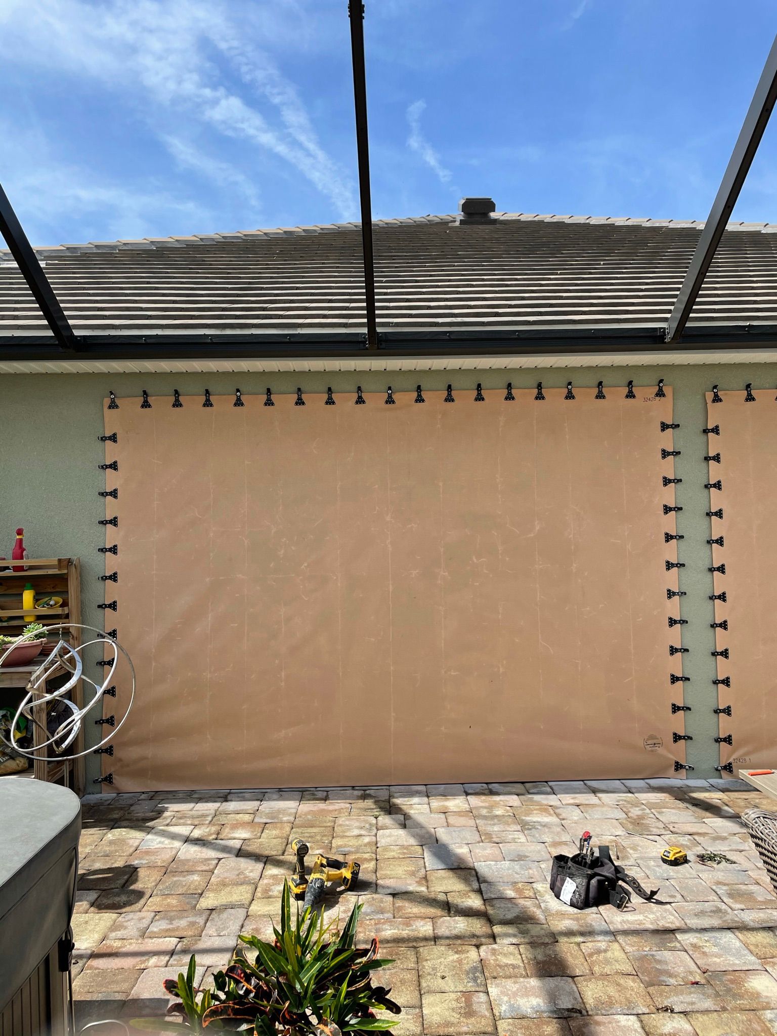 Beige outdoor curtain panel clipped to a patio pergola, with stone paving and a roof in the background.