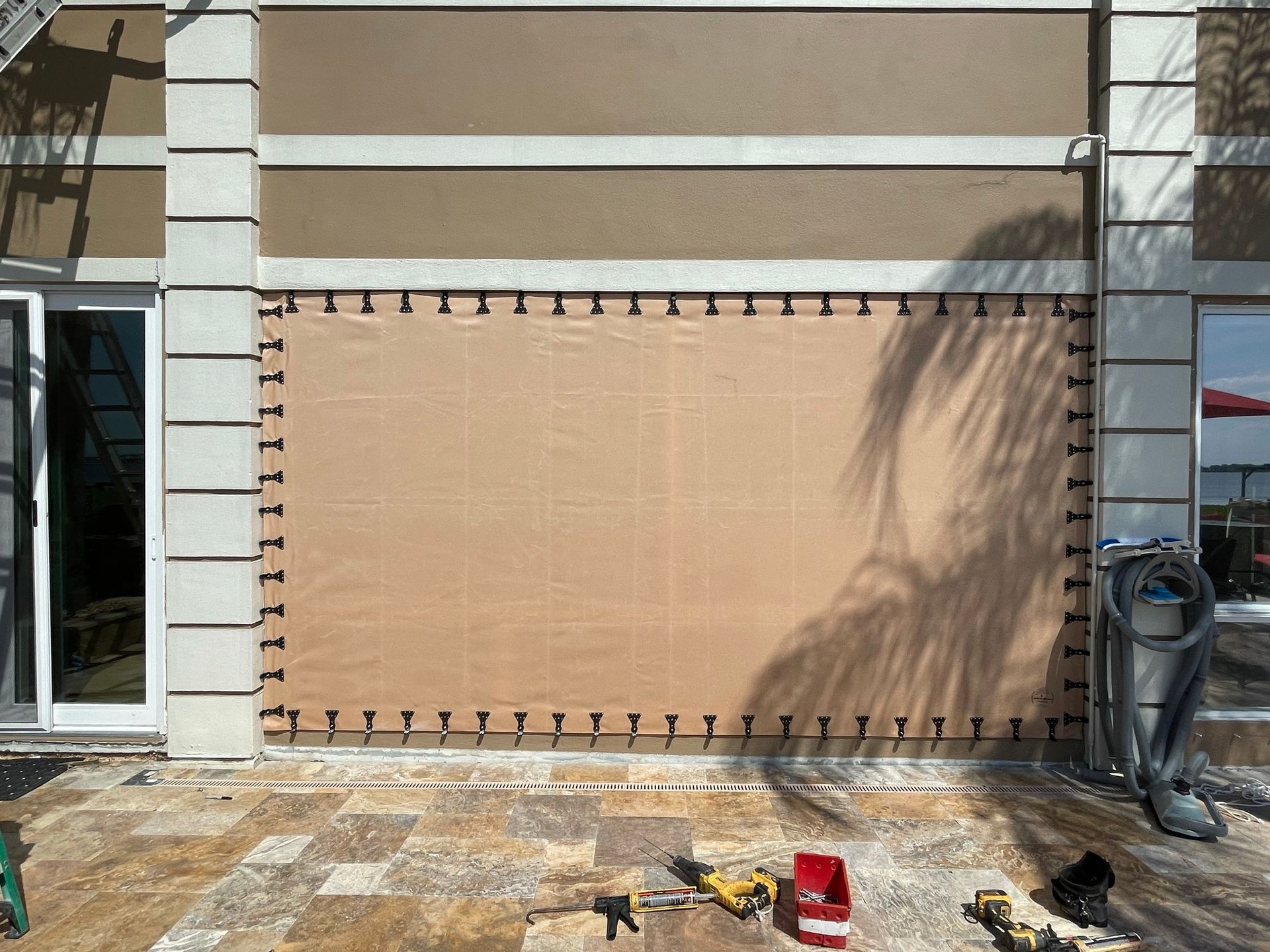 Beige outdoor wall covering with grommets beside patio doors and a coiled hose on the right.