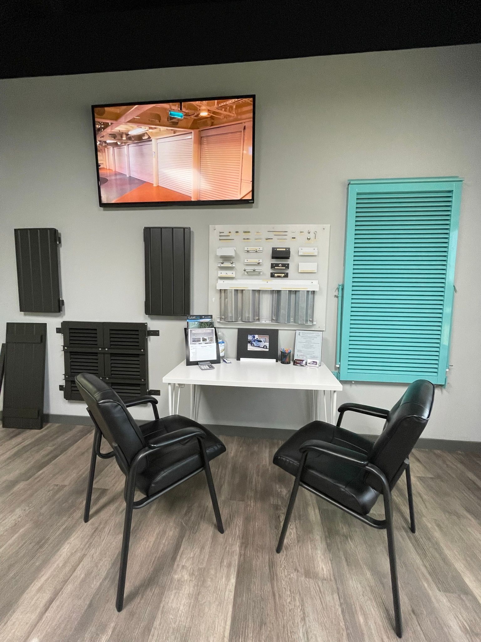 Small waiting area with two black chairs, wall TV, and desk against a gray wall and wood floor.