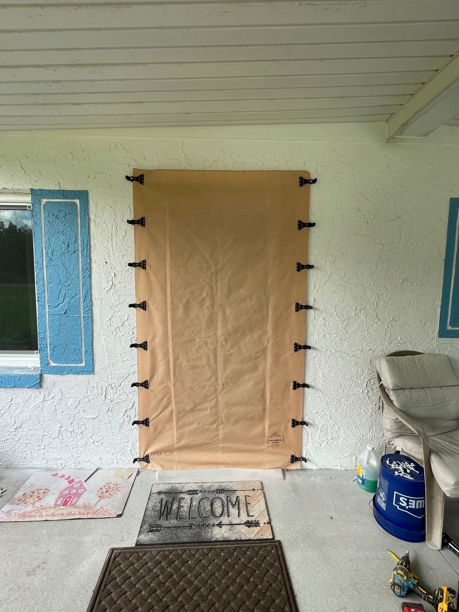 Covered doorway with tan tarp and black clips on a porch between blue-trimmed windows