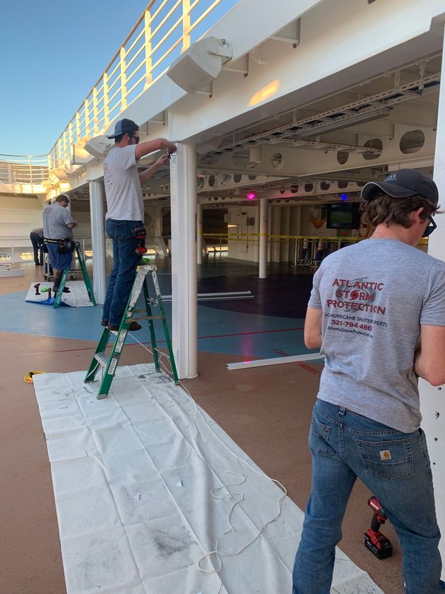 Workers painting an outdoor deck under a covered walkway, with ladders, drop cloths, and tools.