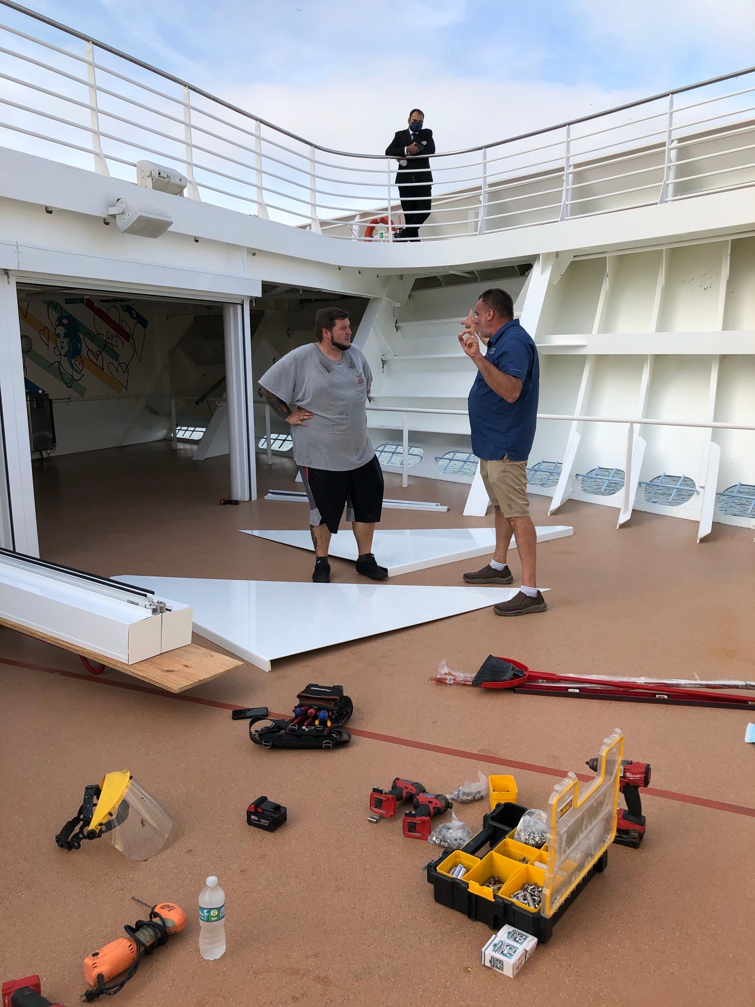 Two men talking on a ship deck with tools scattered on the floor and another person on the upper level.