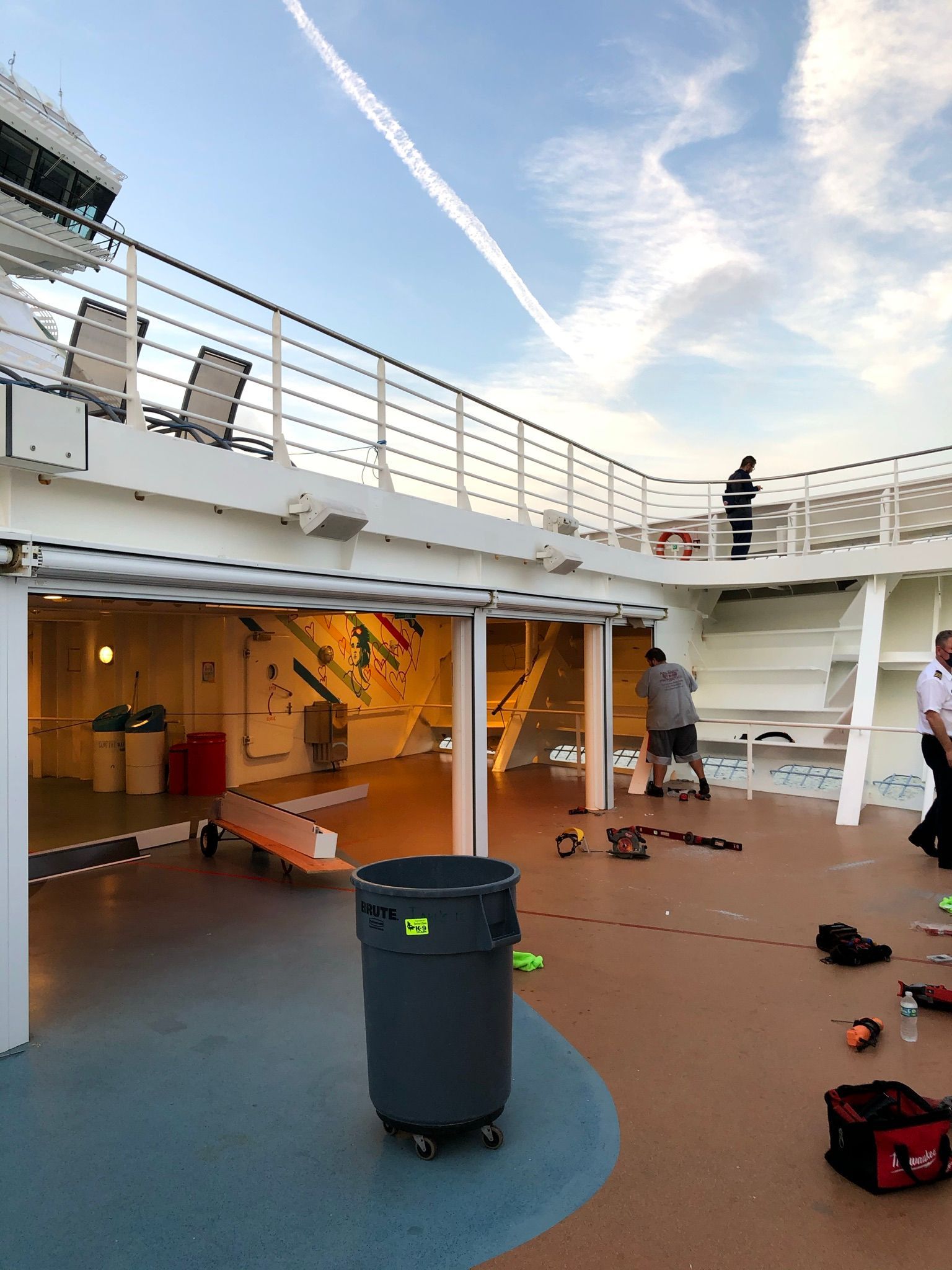 Ship deck with scattered shoes, a trash bin, and people leaning on the upper railing under a blue sky.