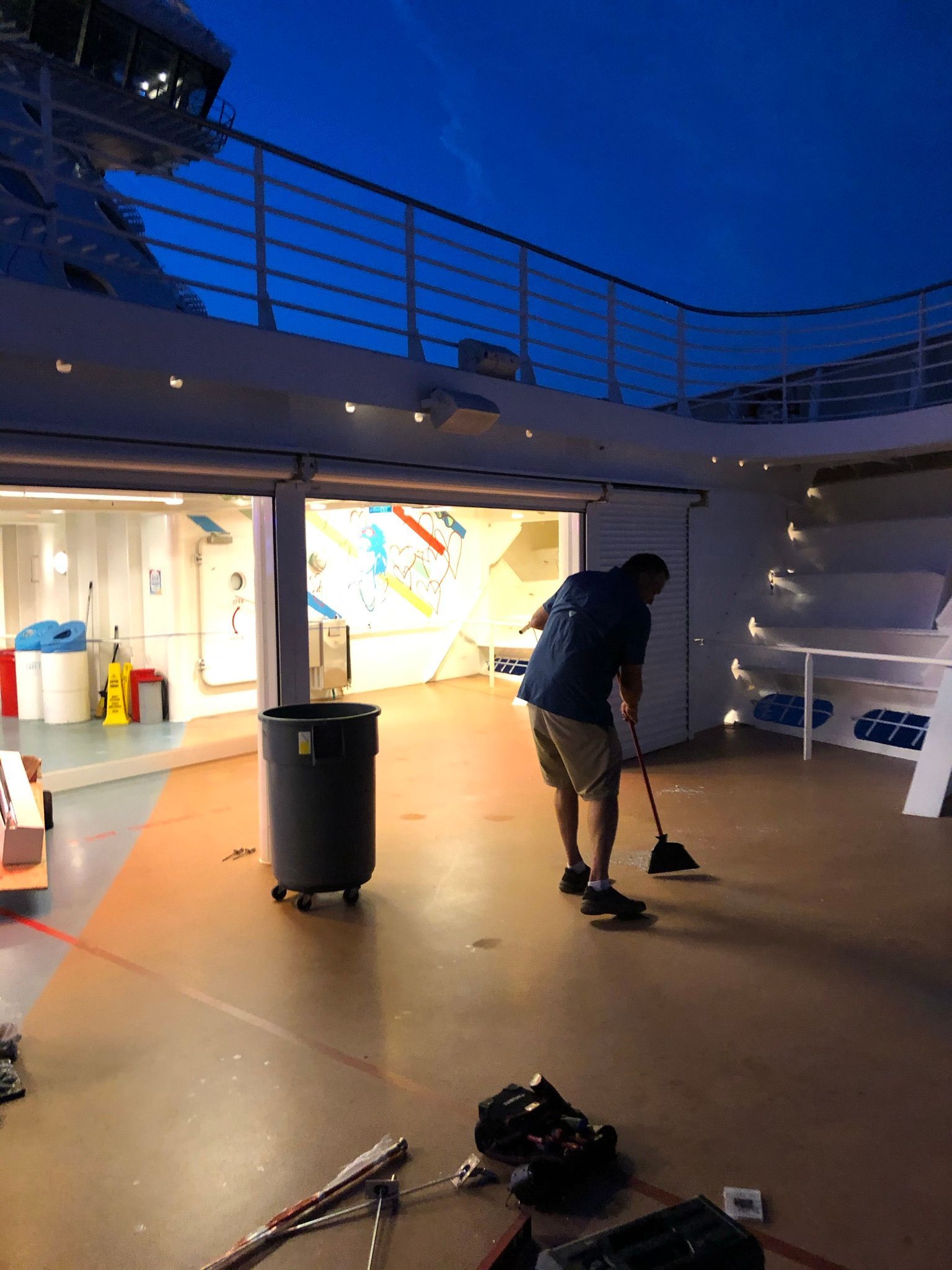 Person sweeping deck at dusk on an outdoor boat or rooftop with blue sky and lit doorway