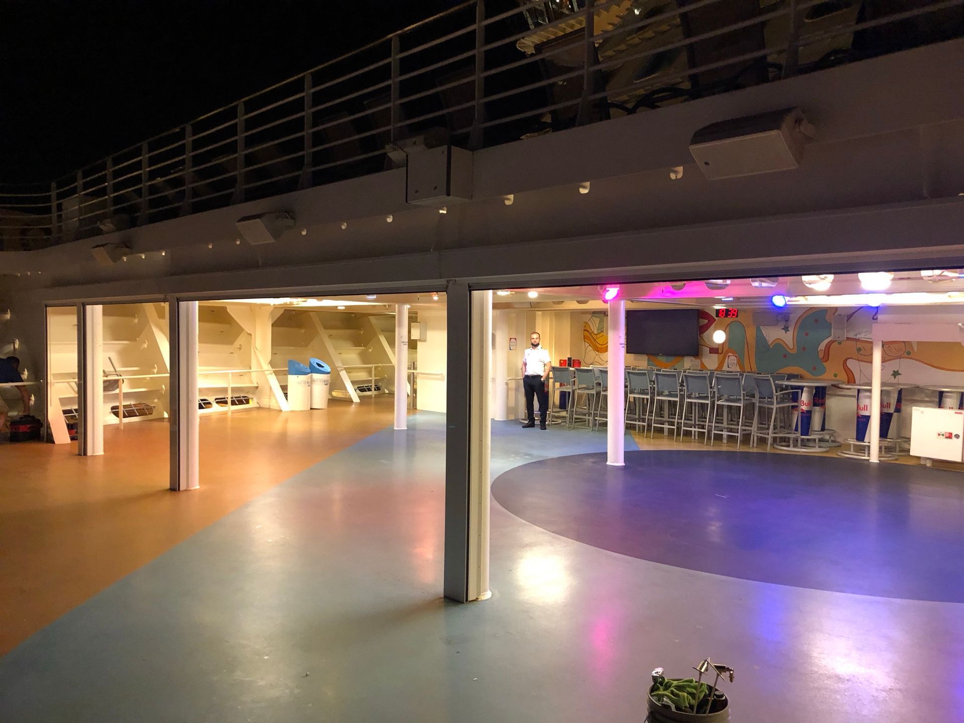 Open indoor deck with colorful floor lighting, bar stools, and a few people in a lounge area at night