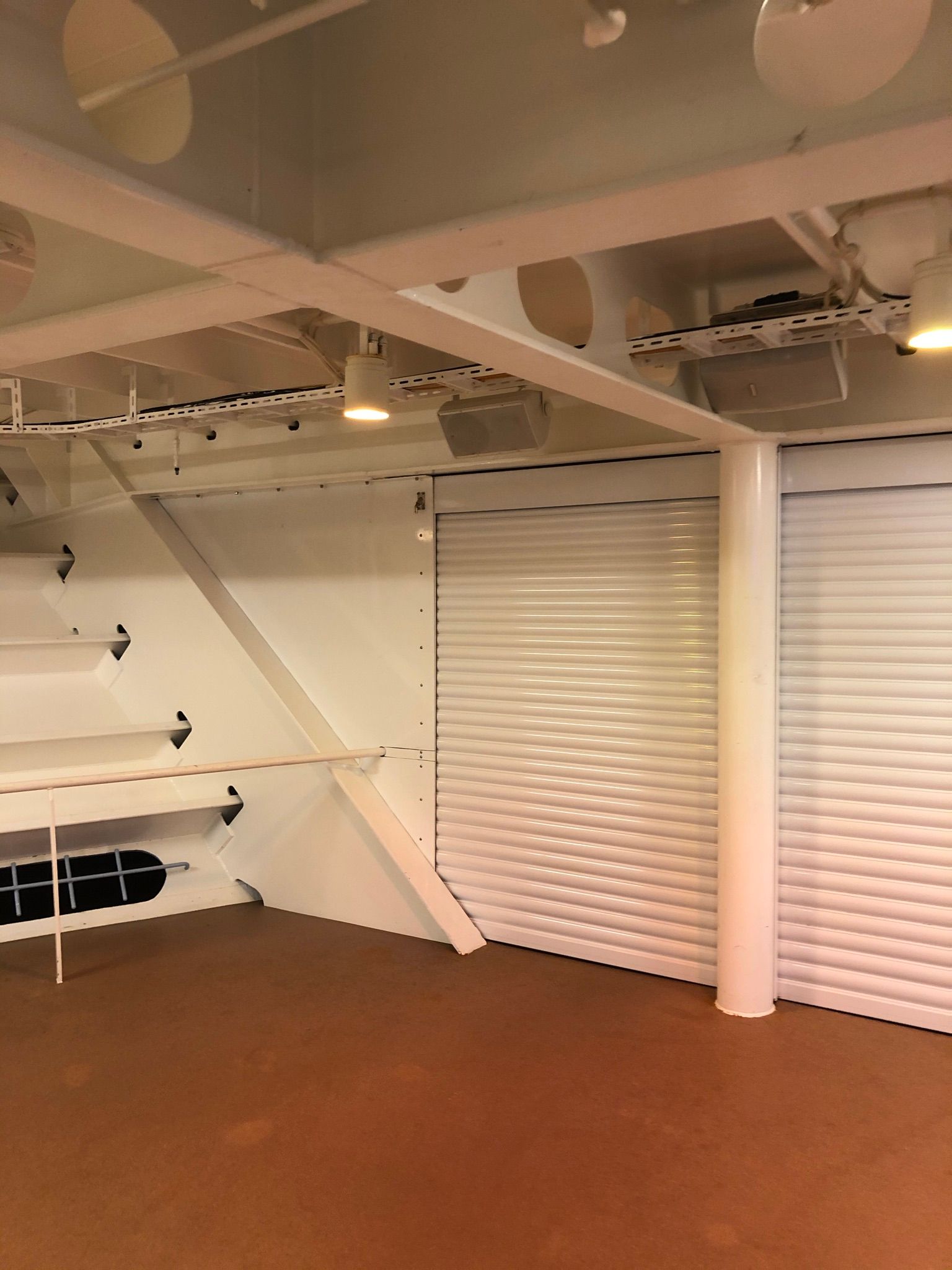 Empty indoor loft with white metal beams, staircase, and beige roller shutters under warm lighting