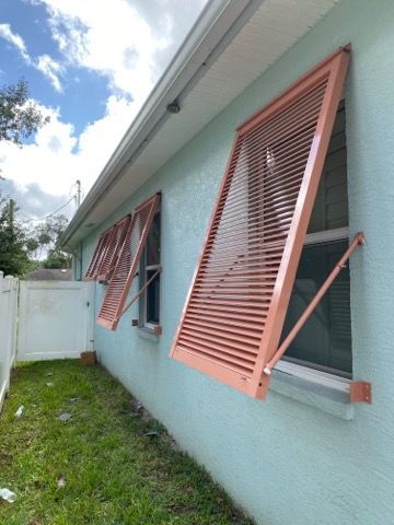 Open windows with coral shutters on a light blue house beside a grassy yard and white fence