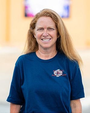 Woman in a blue T-shirt smiling indoors against a softly blurred background.
