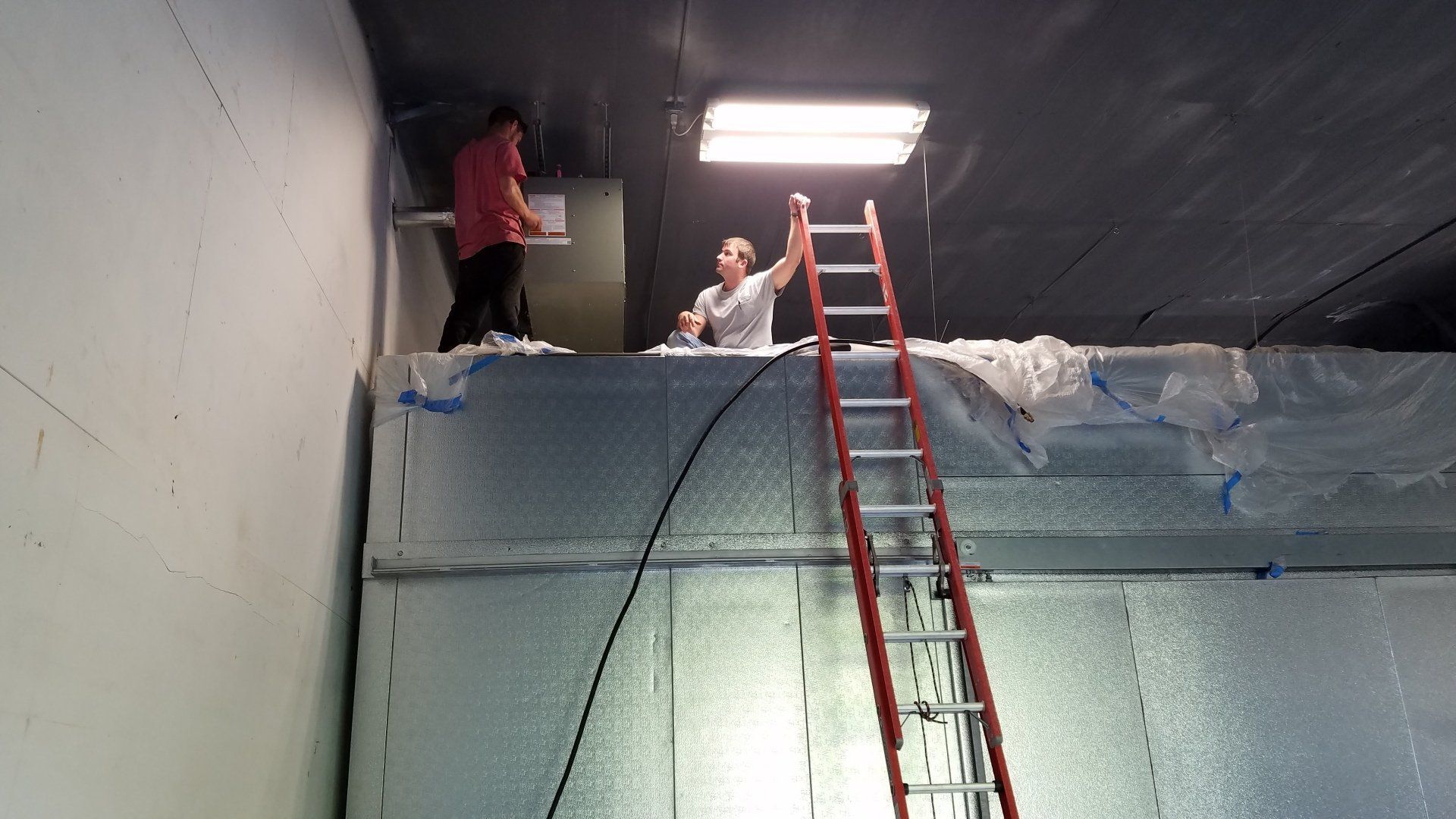 Two people work inside a room, one on a ladder, the other on a ledge; construction.