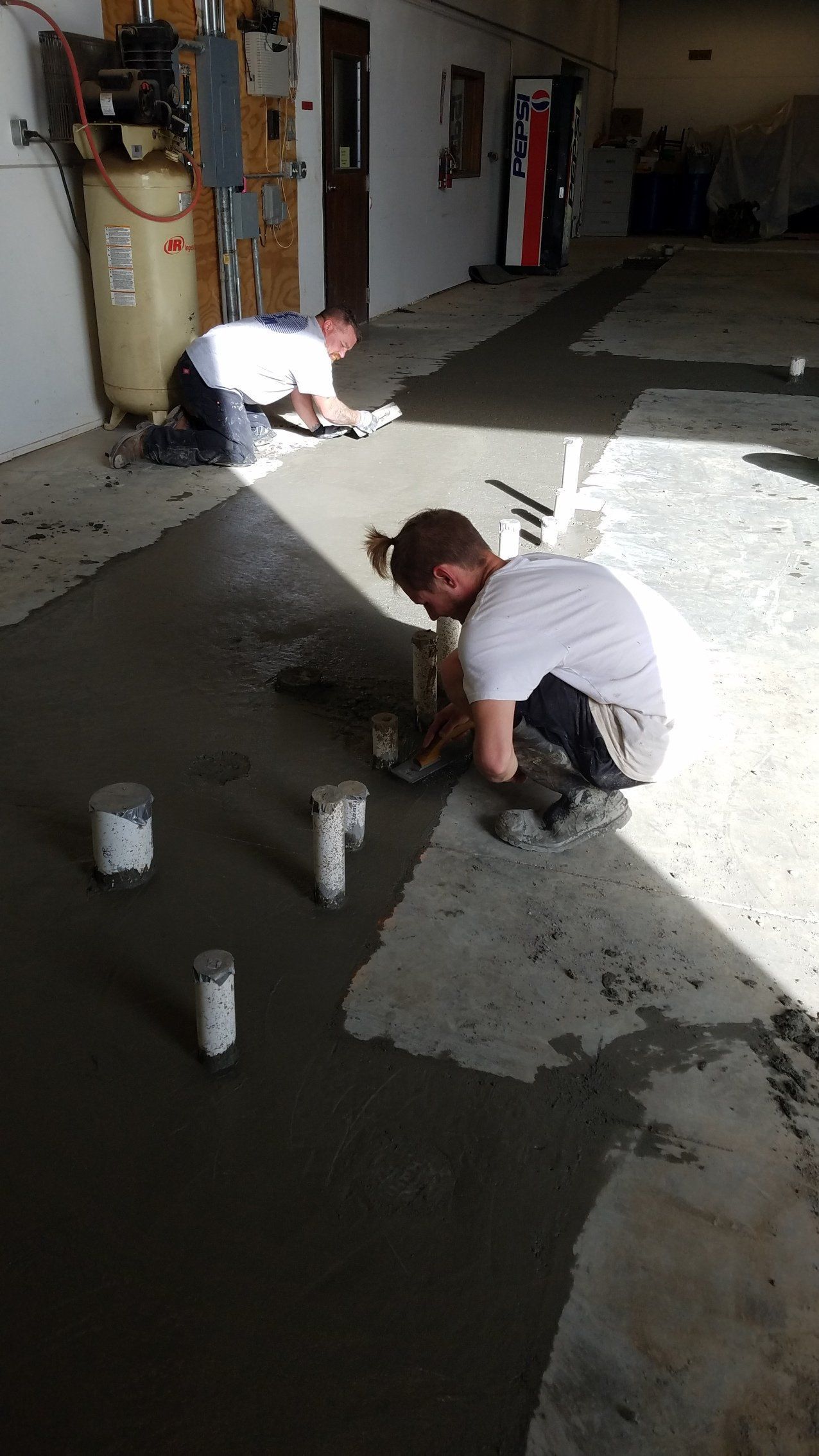 Two people smoothing wet concrete on a garage floor. Cylinders of concrete are in the foreground.