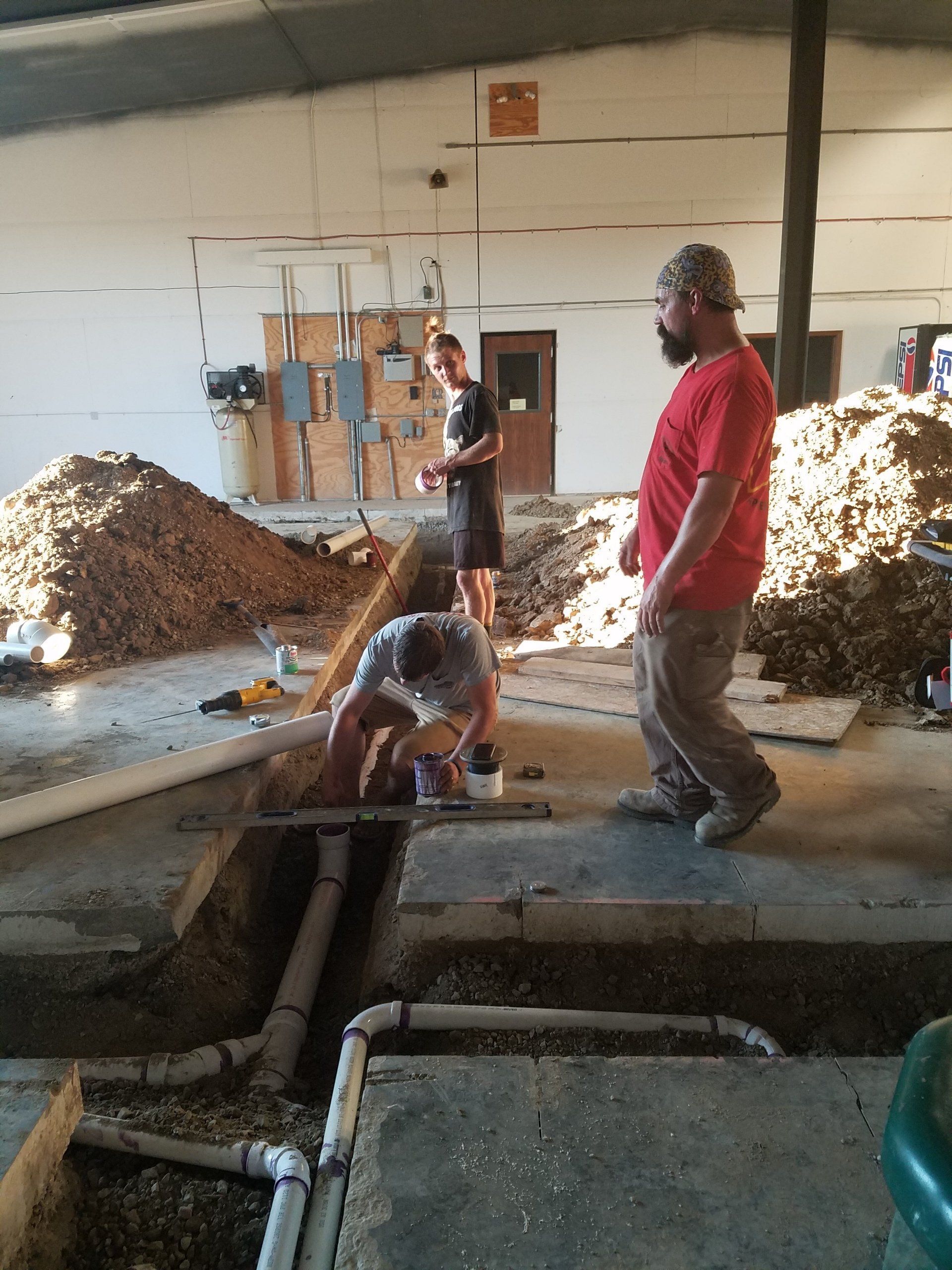 Men working on plumbing in a building; one is crouched, others standing. Gravel and dirt are visible.