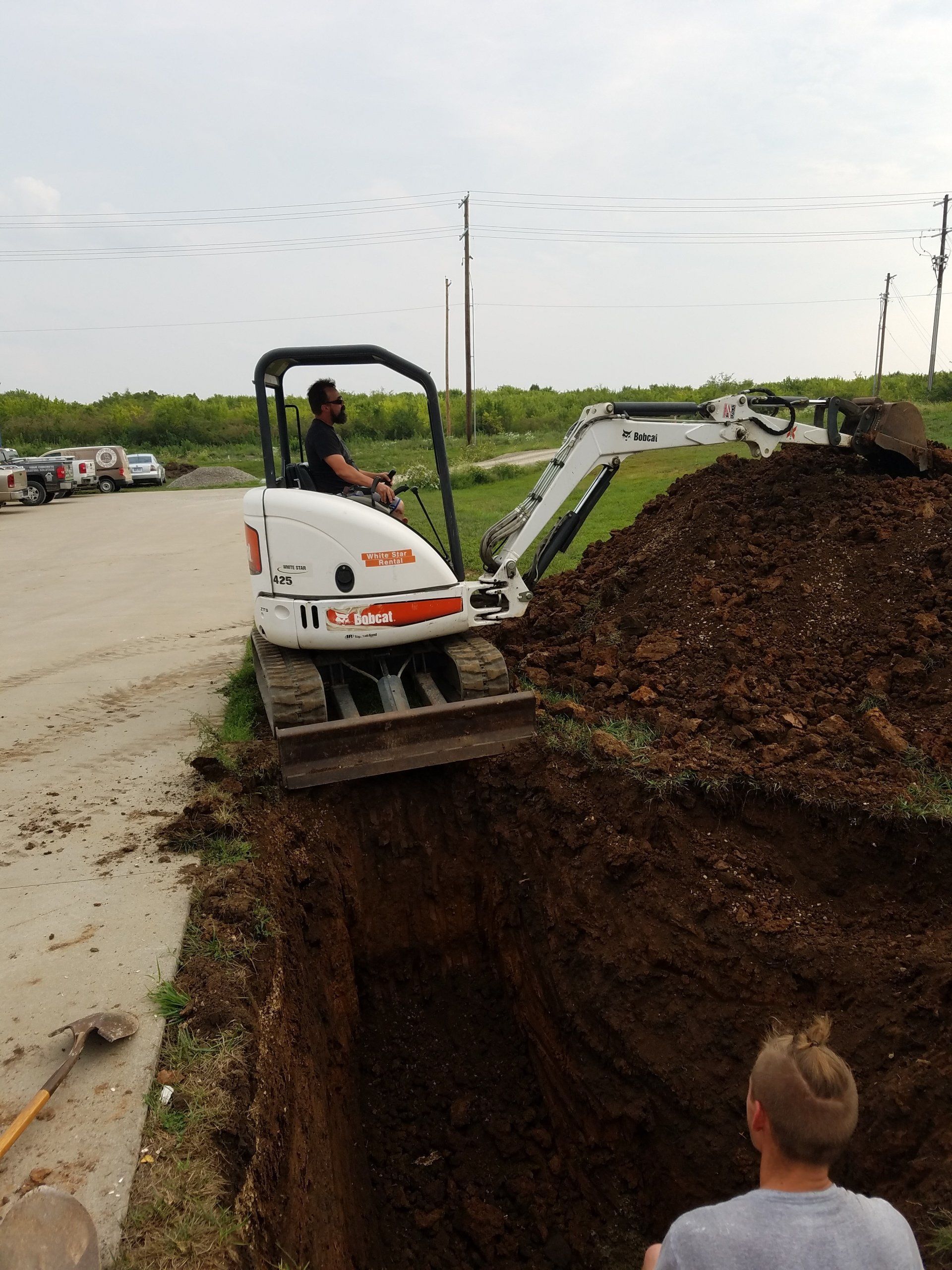 A small excavator digging a trench, with a man operating it, another man watching nearby. Outdoors.