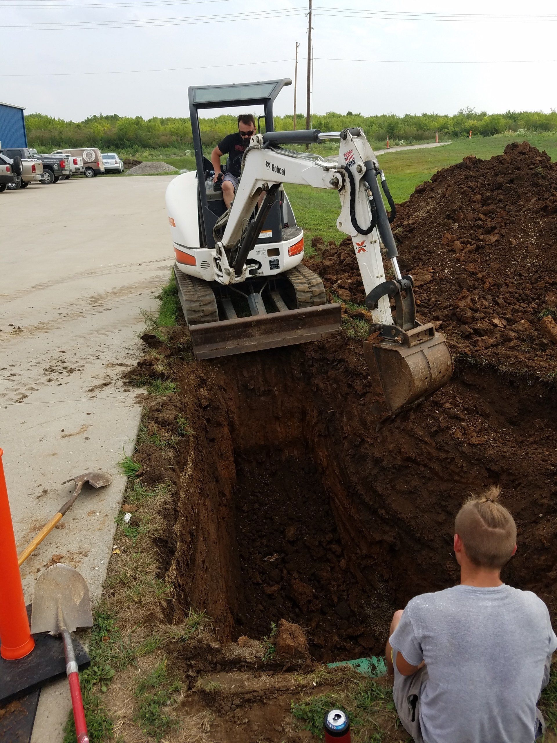Man operating an excavator digging a trench, another man observing. Dirt pile, road, orange cone.