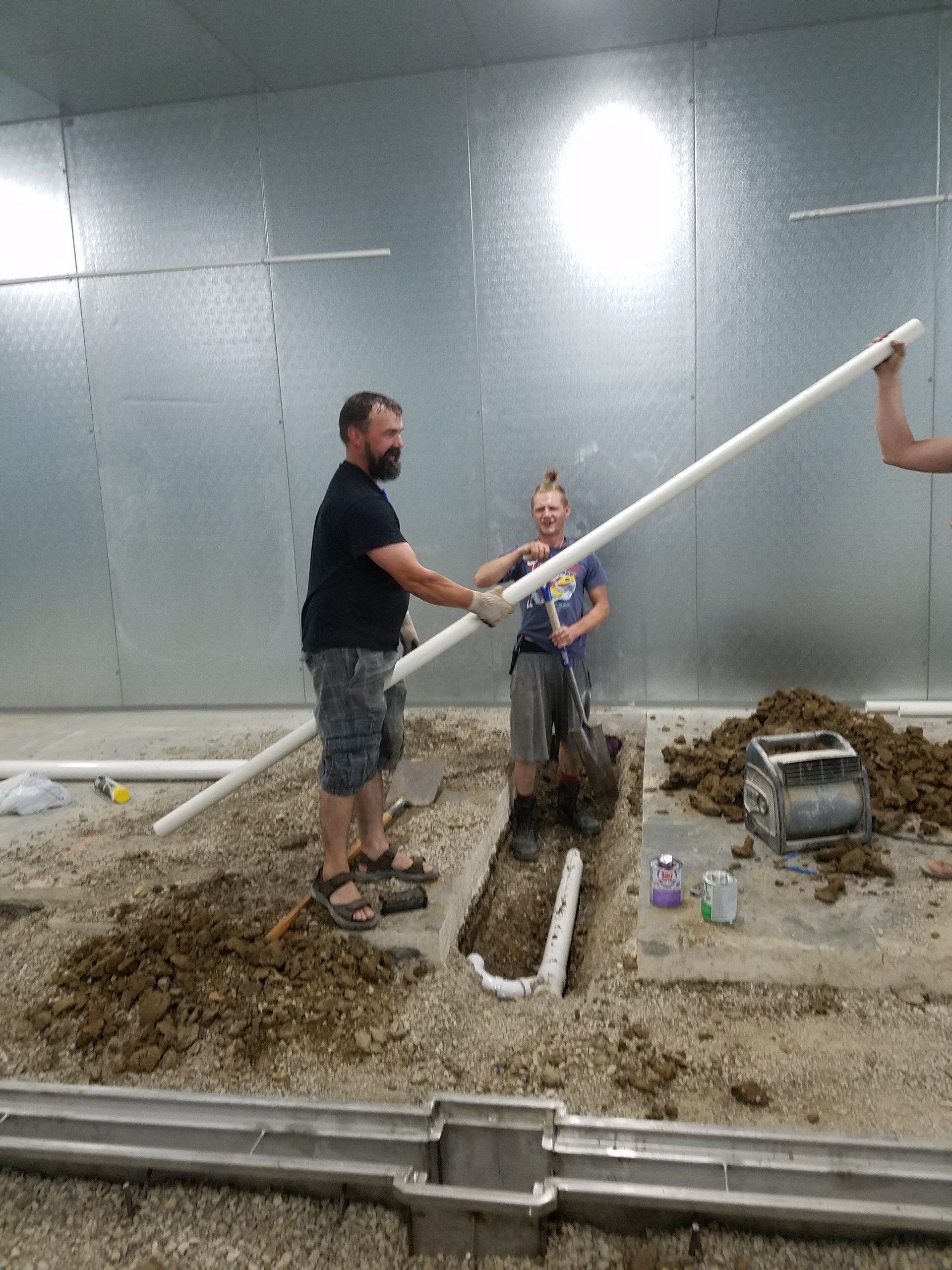 Workers installing PVC pipes in a trench inside a building. One man holds a long pipe.