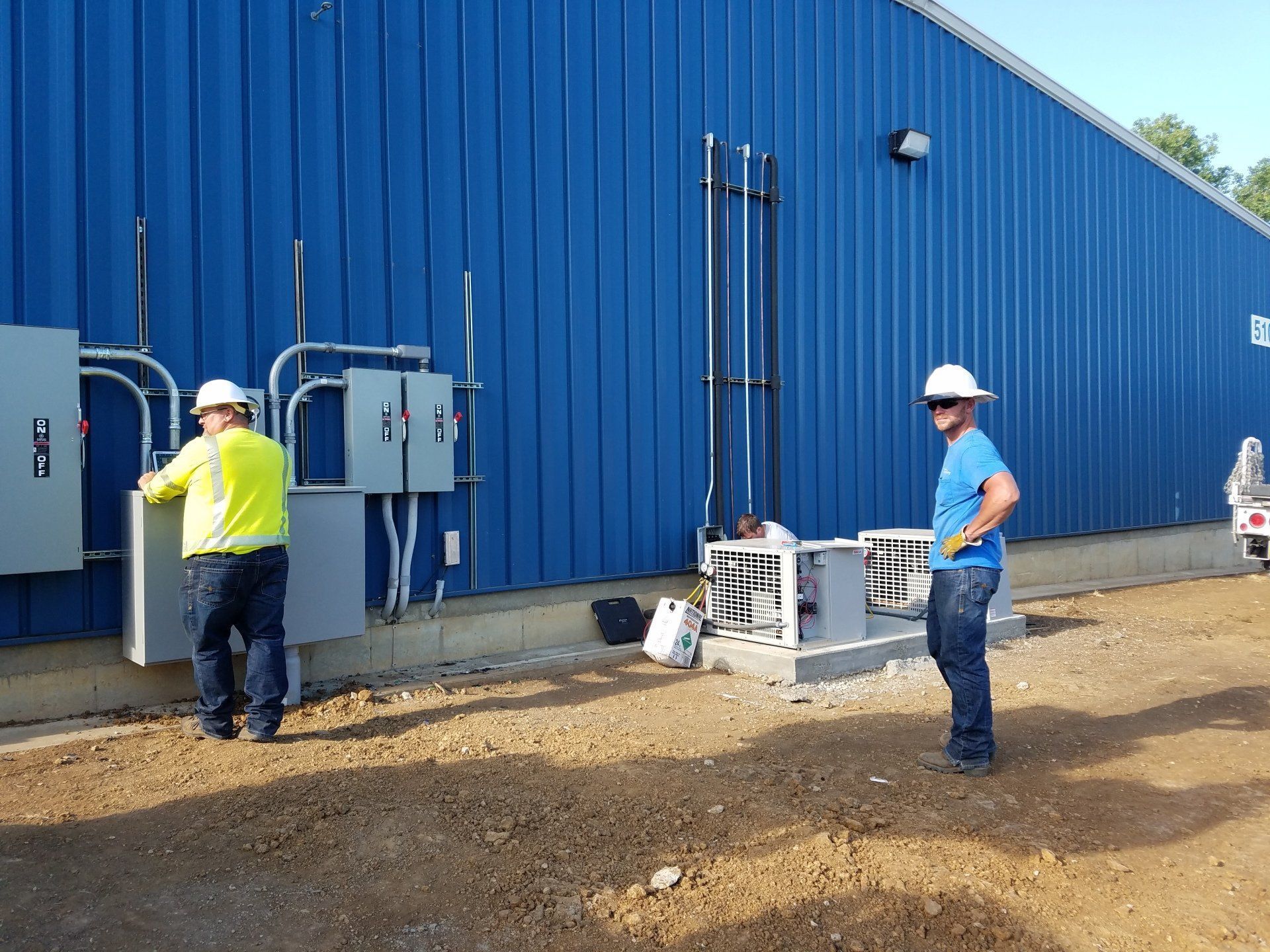Two workers near blue industrial building, one in yellow vest opening a gray electrical panel, the other in blue shirt, hands on hip.