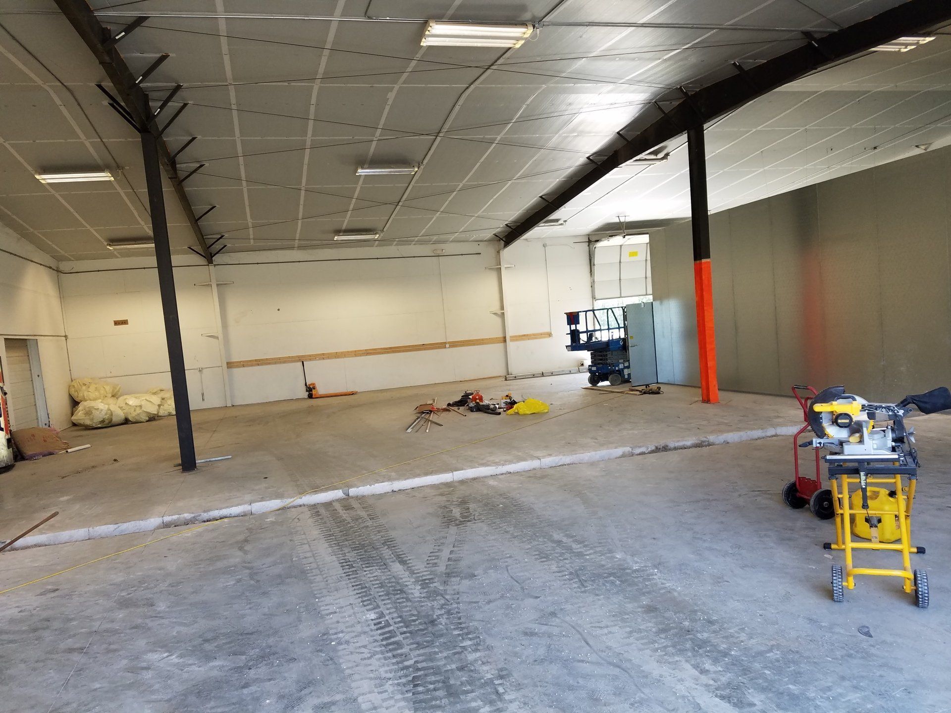 Empty warehouse interior, concrete floor, white walls, support beams. Construction tools and materials scattered.