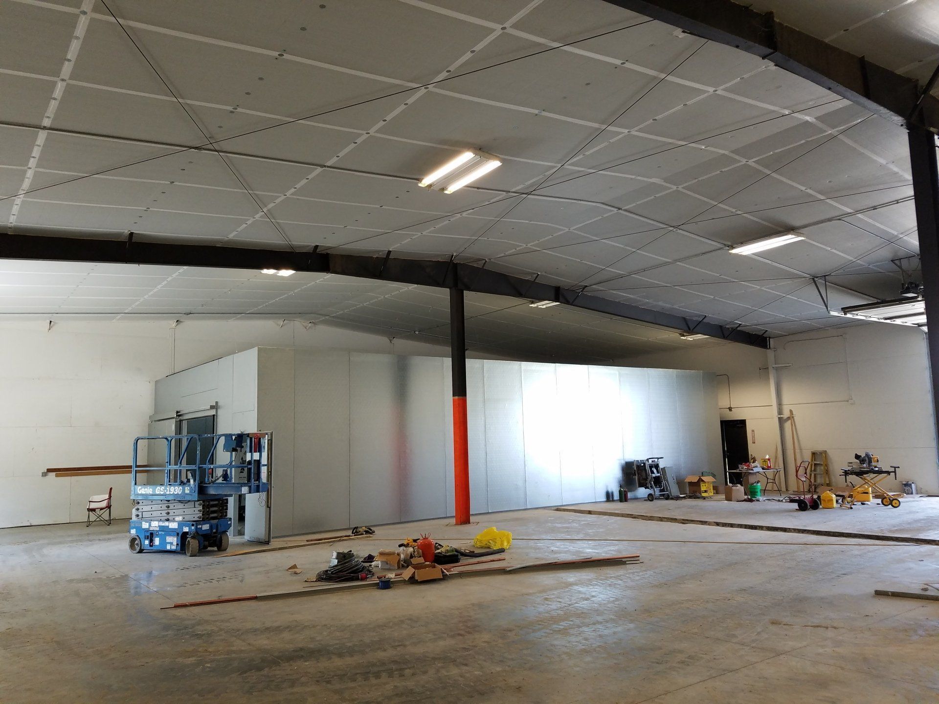 Interior of a large, empty warehouse. A lift, construction materials, and white walls are visible.