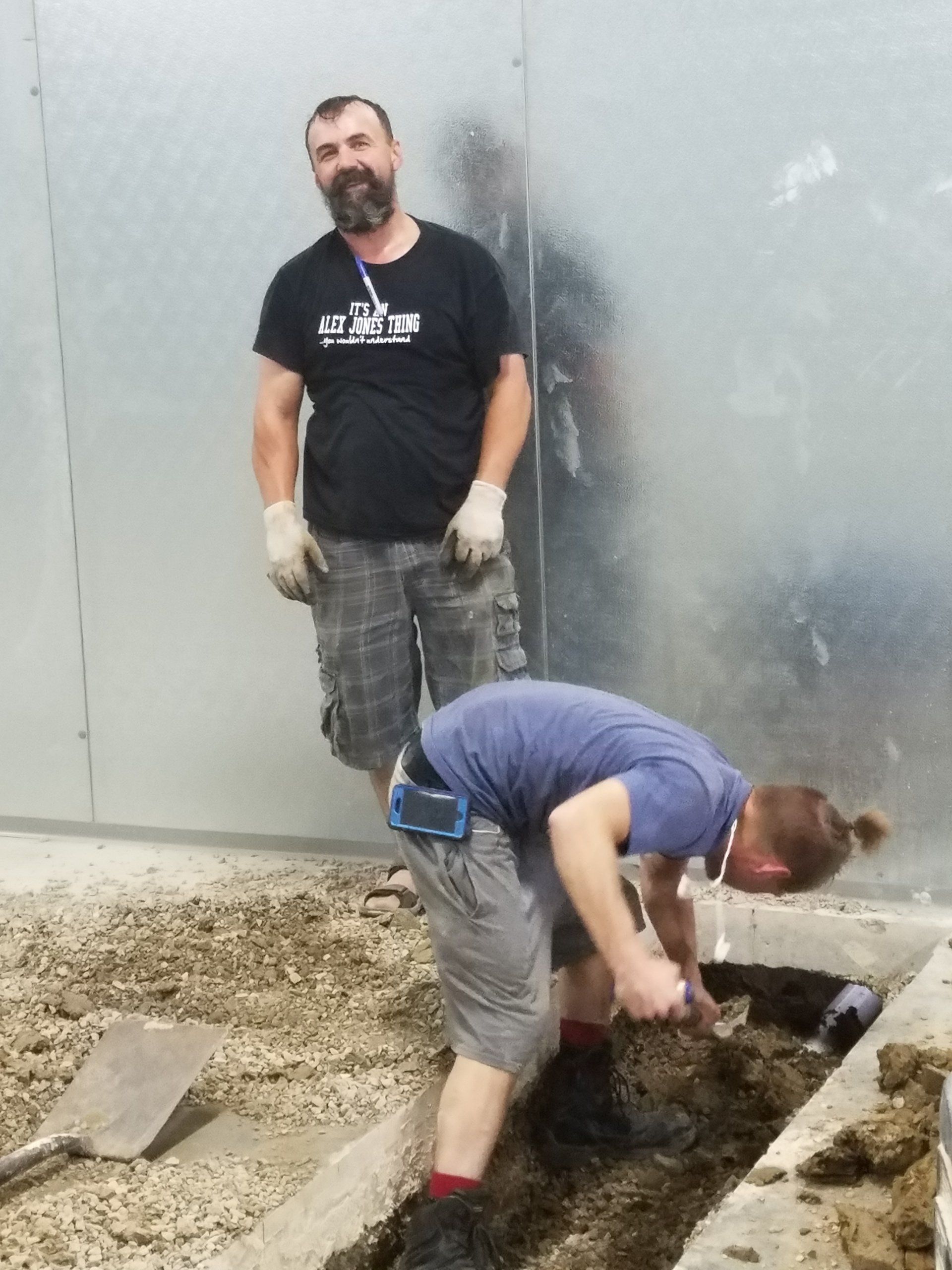 Man watches as another digs in a trench. Both wear work clothes. Concrete wall.