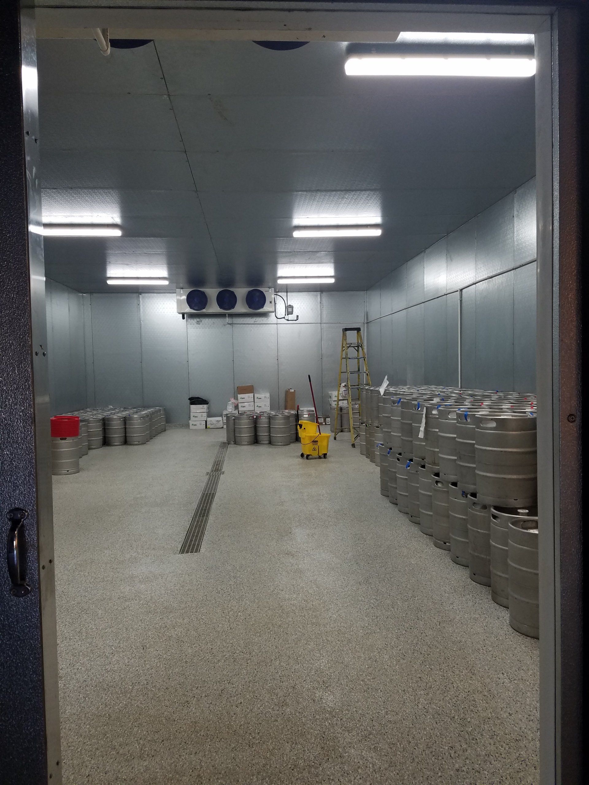 Walk-in cooler, filled with stacks of silver beer kegs. White walls, gray speckled floor, overhead lights.