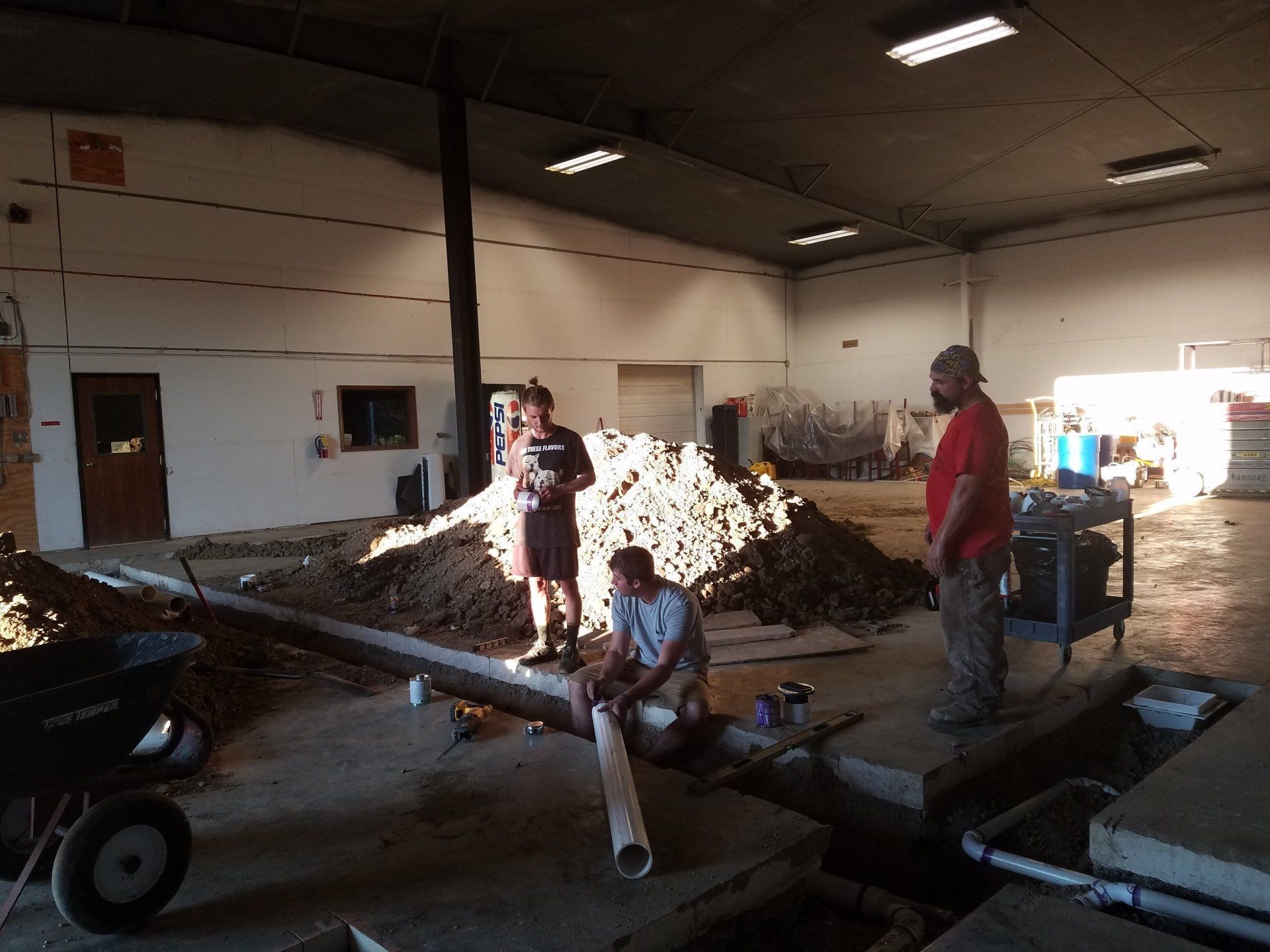 Three people working in an industrial building with dirt pile and concrete trenches.
