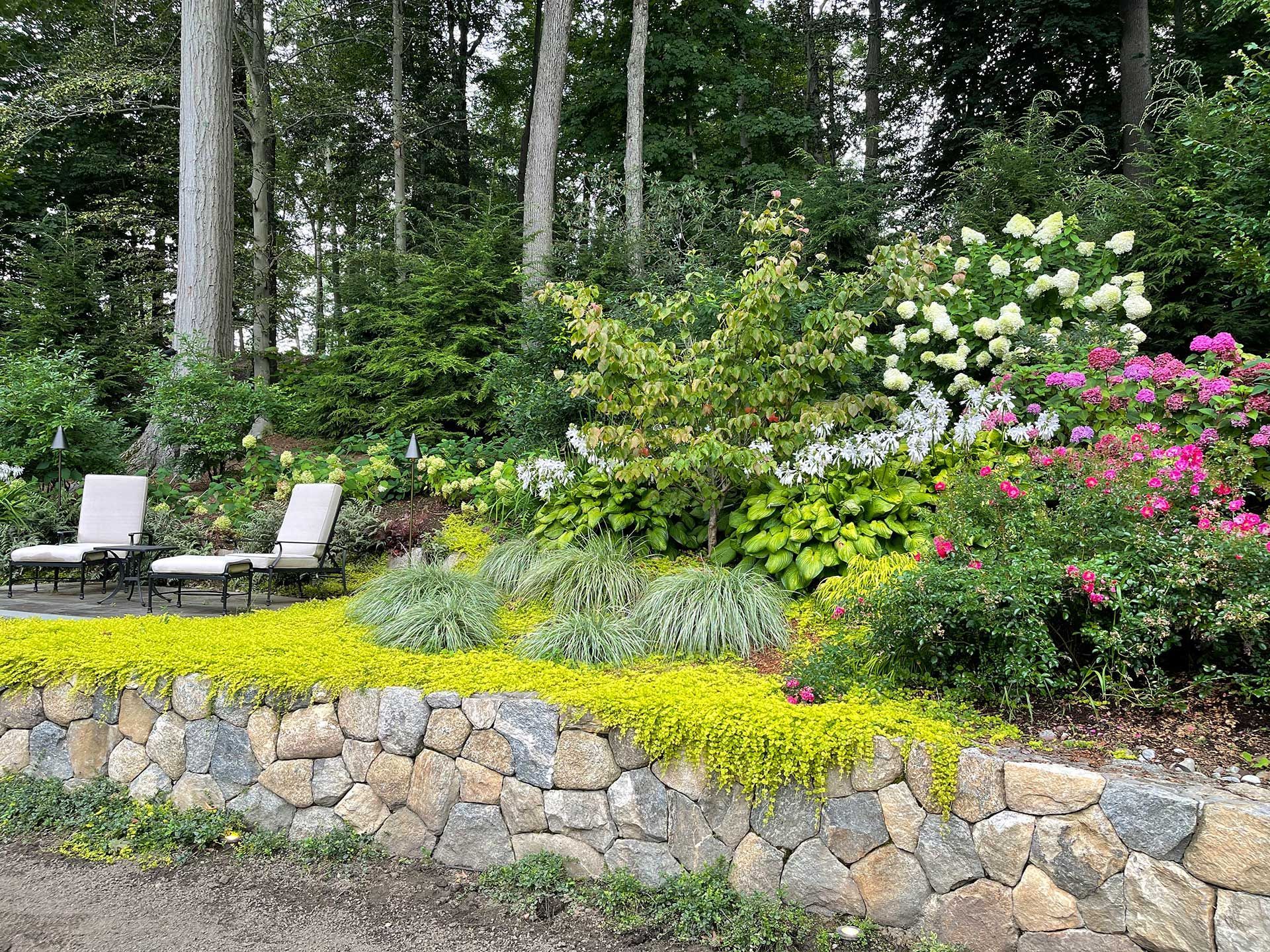 a stone wall surrounds a lush green garden with chairs and flowers