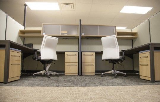 Empty office cubicles with two white swivel chairs and desks under fluorescent lights.