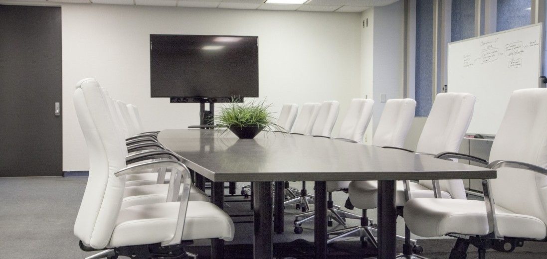 Modern conference room with a long table, white chairs, wall-mounted TV, and windows on the right.