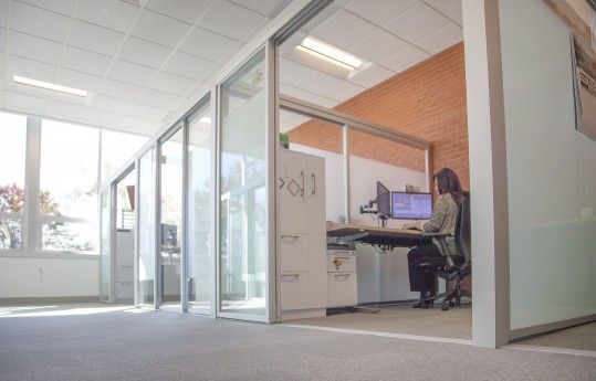 Bright office hallway with glass-walled cubicles and a person working at a desk on a laptop.
