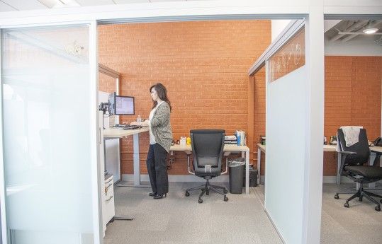 Person standing at a computer in a modern office cubicle with orange brick wall and black chairs.