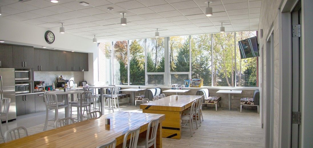 Bright open dining hall with long tables, white chairs, and large windows overlooking trees