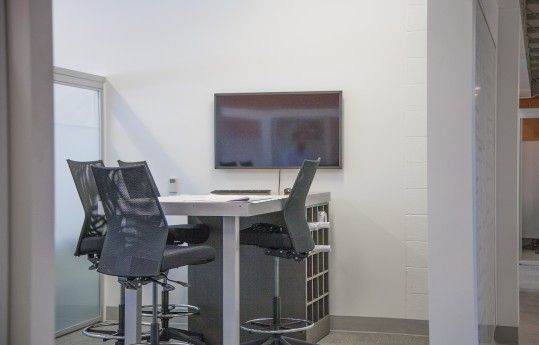 Small office meeting area with chairs around a high table and a wall-mounted TV.