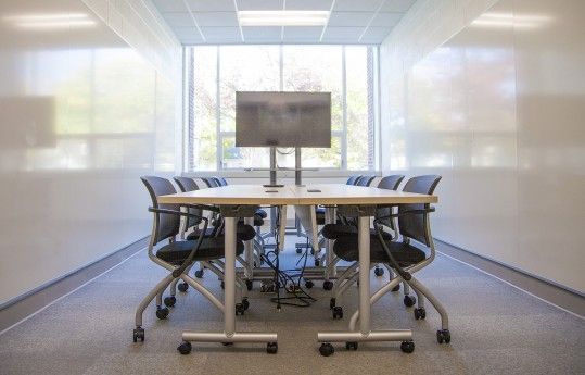 Modern conference room with a long table, rolling chairs, and a monitor at the far end.