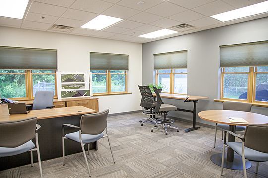 Bright office with desks, chairs, large windows, and gray carpet under fluorescent lights