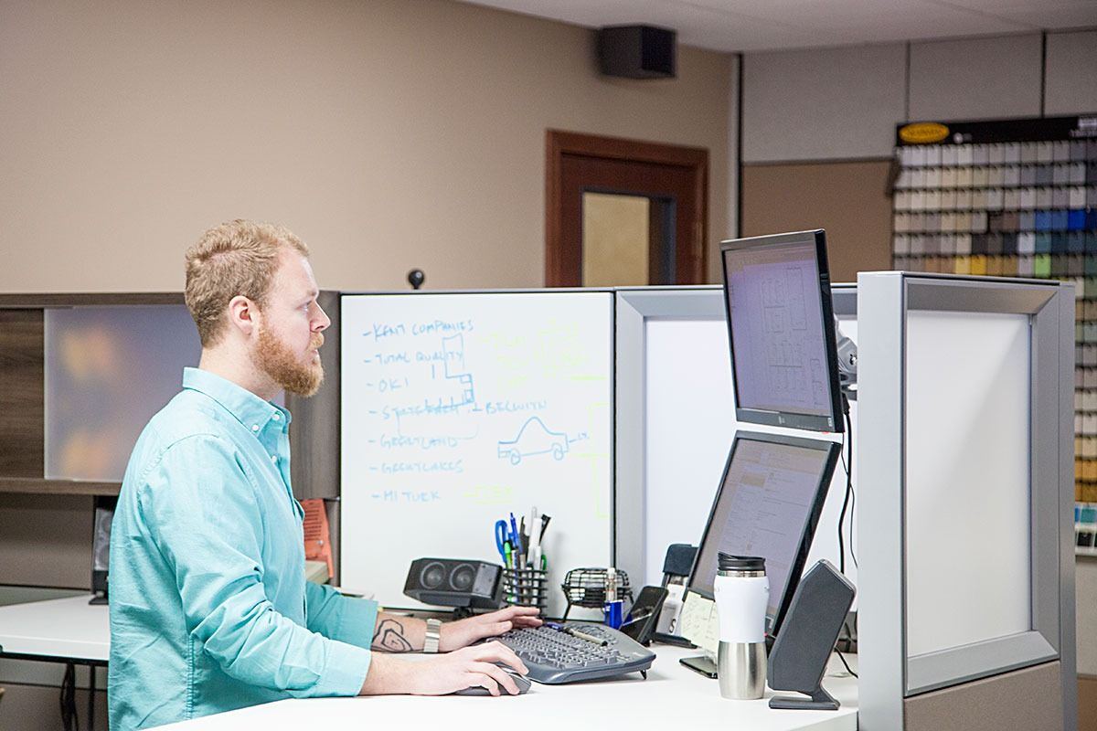 Man working at a cubicle desk with two monitors and a whiteboard in an office
