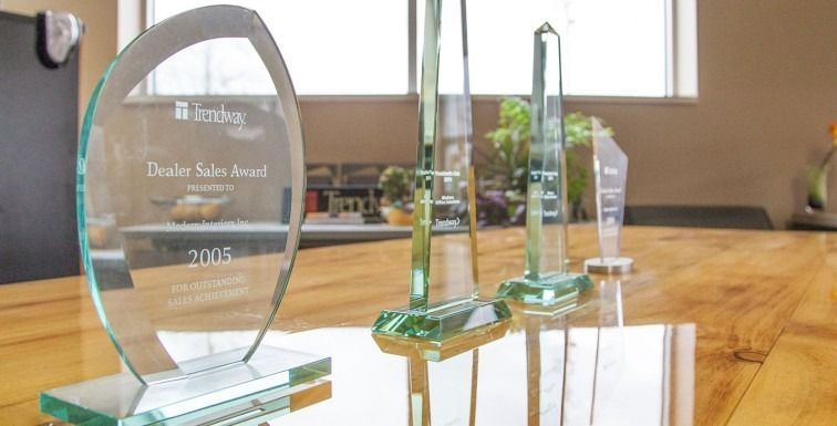 Glass awards on a wooden table in front of a window, including a clear oval trophy and green crystal plaques