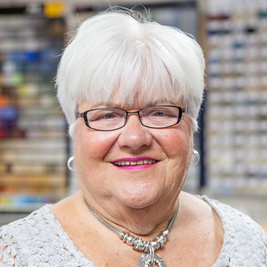 Smiling woman with white hair and glasses, wearing a silver necklace, standing indoors with blurred shelves behind her