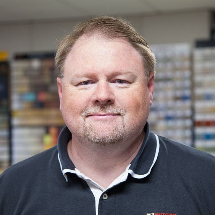 Smiling man in a navy polo shirt standing indoors with shelves blurred in the background