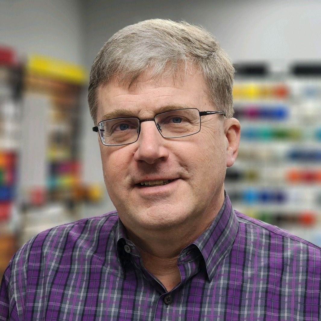 Man wearing glasses and a purple plaid shirt in a store aisle with blurred shelves behind him