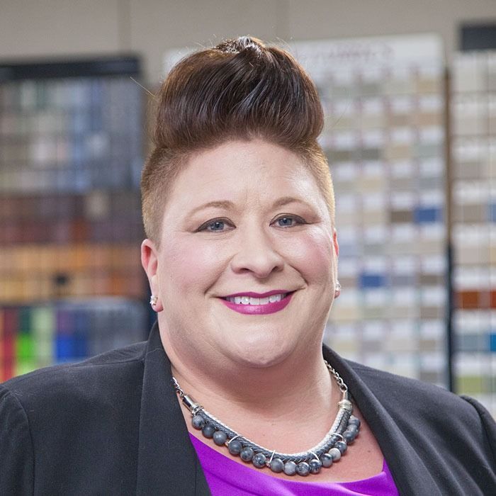 Smiling person in a black blazer and purple top, wearing a statement necklace, seated in front of shelves.