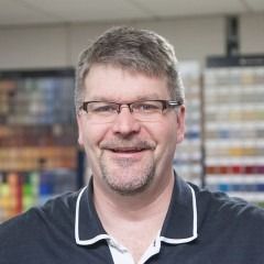 Smiling man with glasses in a dark polo shirt, standing in front of shelves of paint samples.