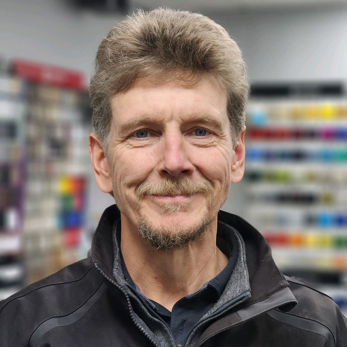 Portrait of a smiling man in a black jacket, standing in front of blurred store shelves