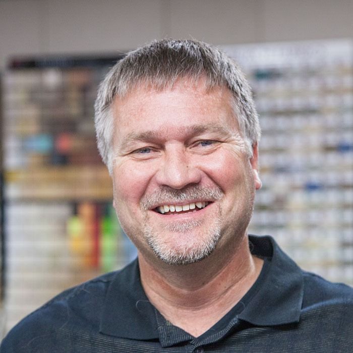 Smiling man with gray hair and beard in a dark polo shirt, standing indoors with blurred shelves behind him