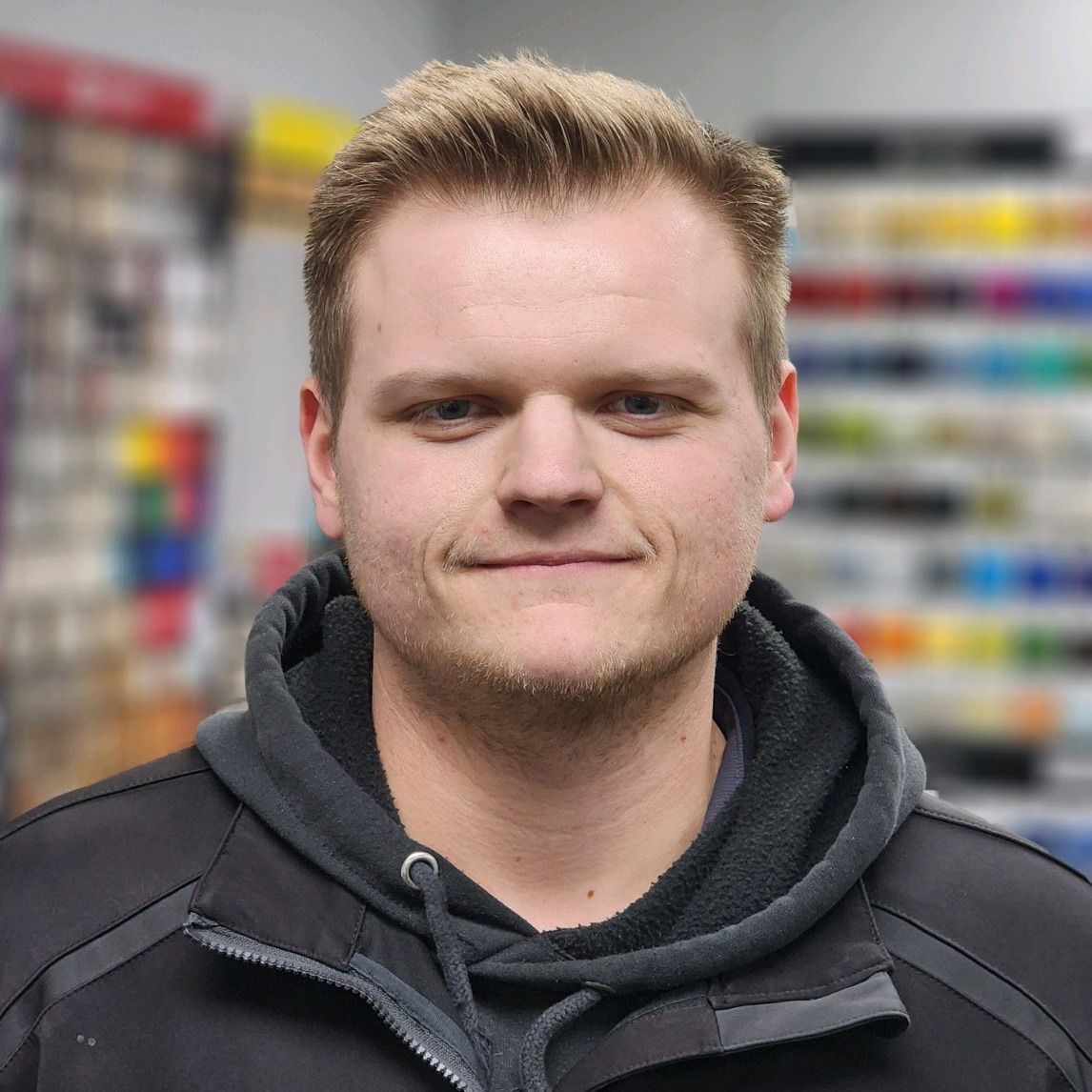 Portrait of a smiling person in a dark jacket, standing in a store aisle with blurred shelves behind.