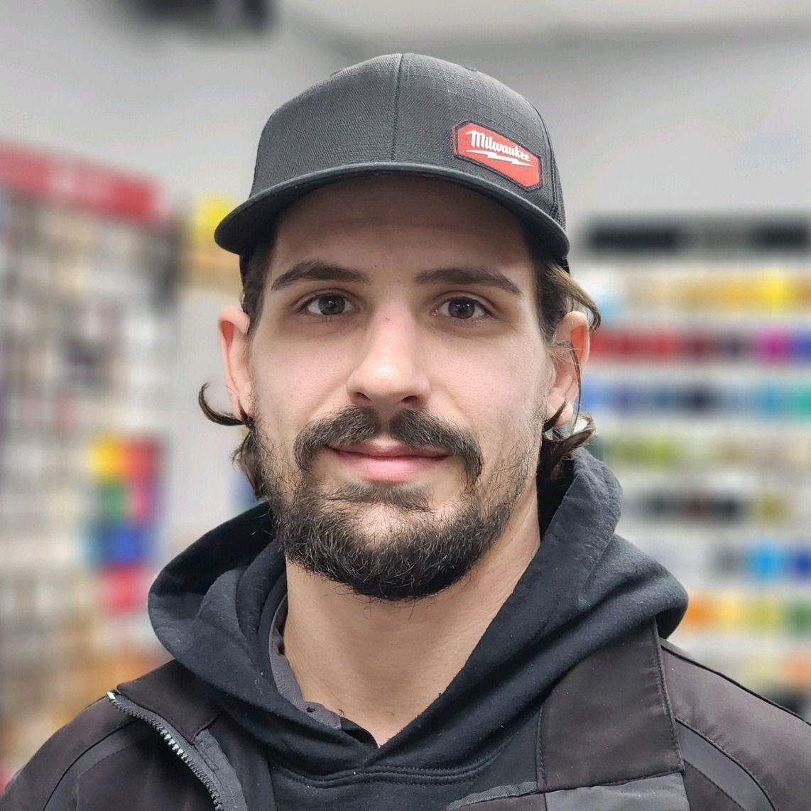 Man in a black cap and hoodie, standing in a store aisle with blurred shelves behind him.