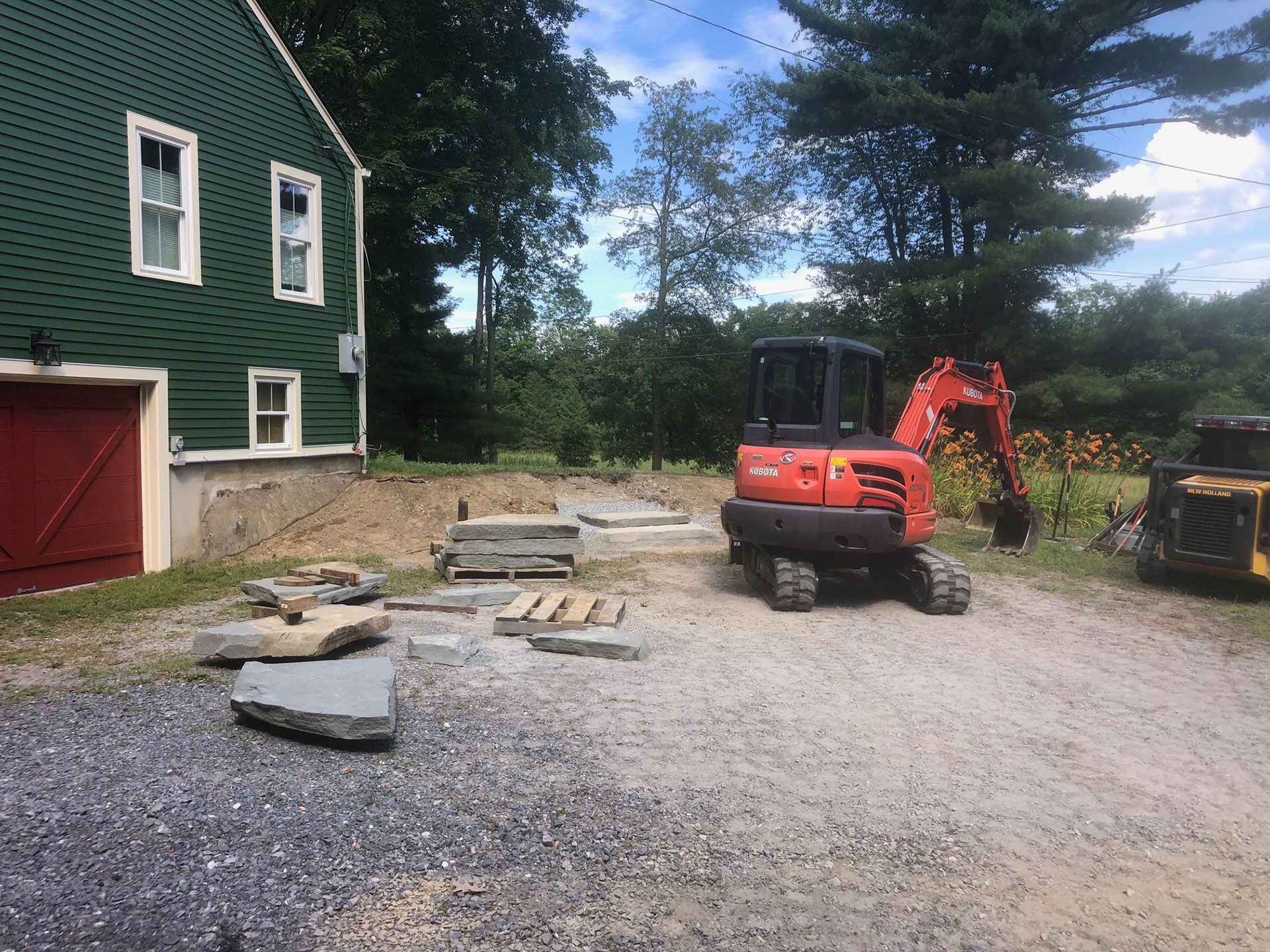 A small orange excavator on a gravel lot next to a green building. Construction supplies are nearby.