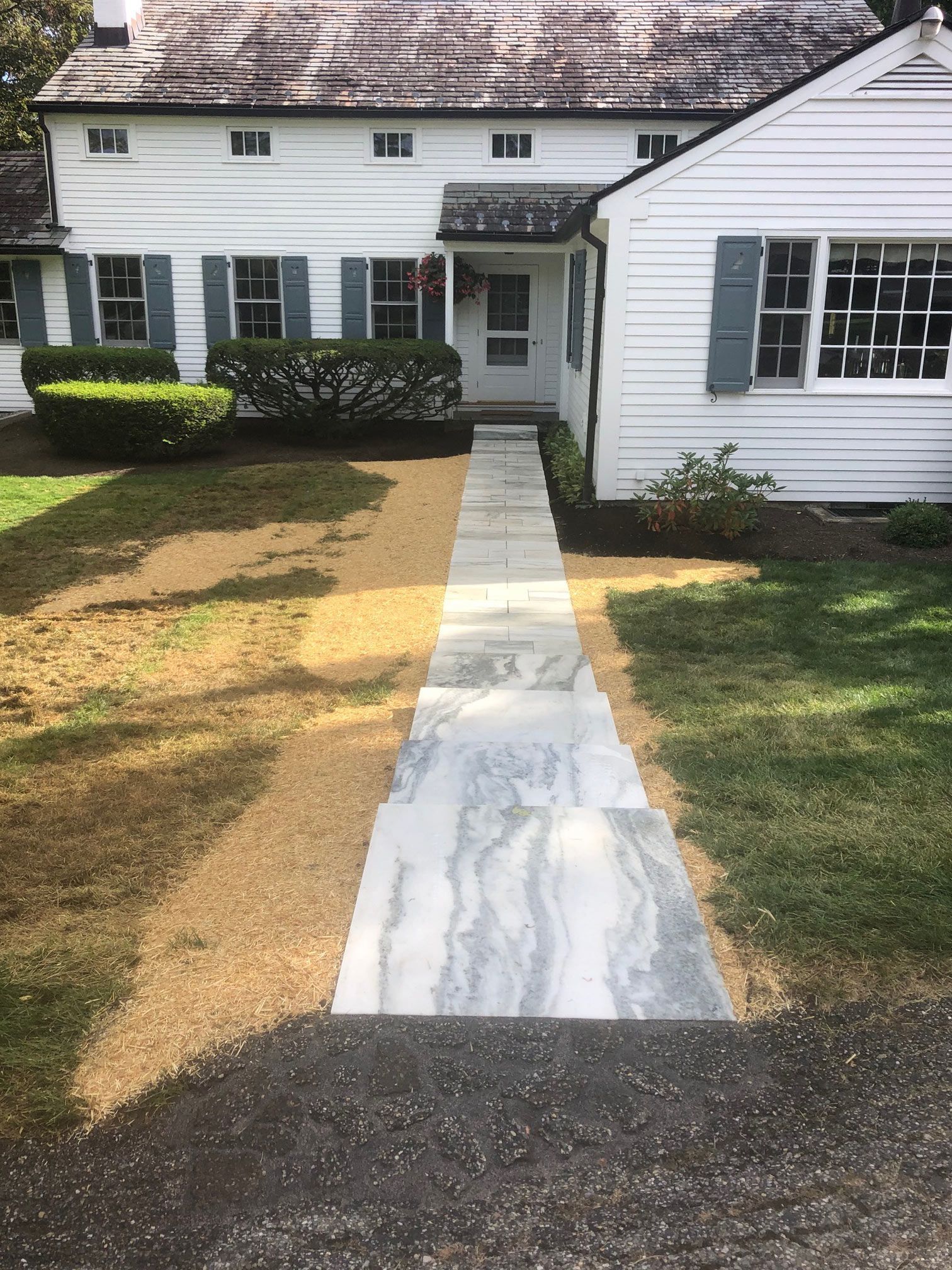 Stone walkway leading to a white house with blue shutters, surrounded by grass.