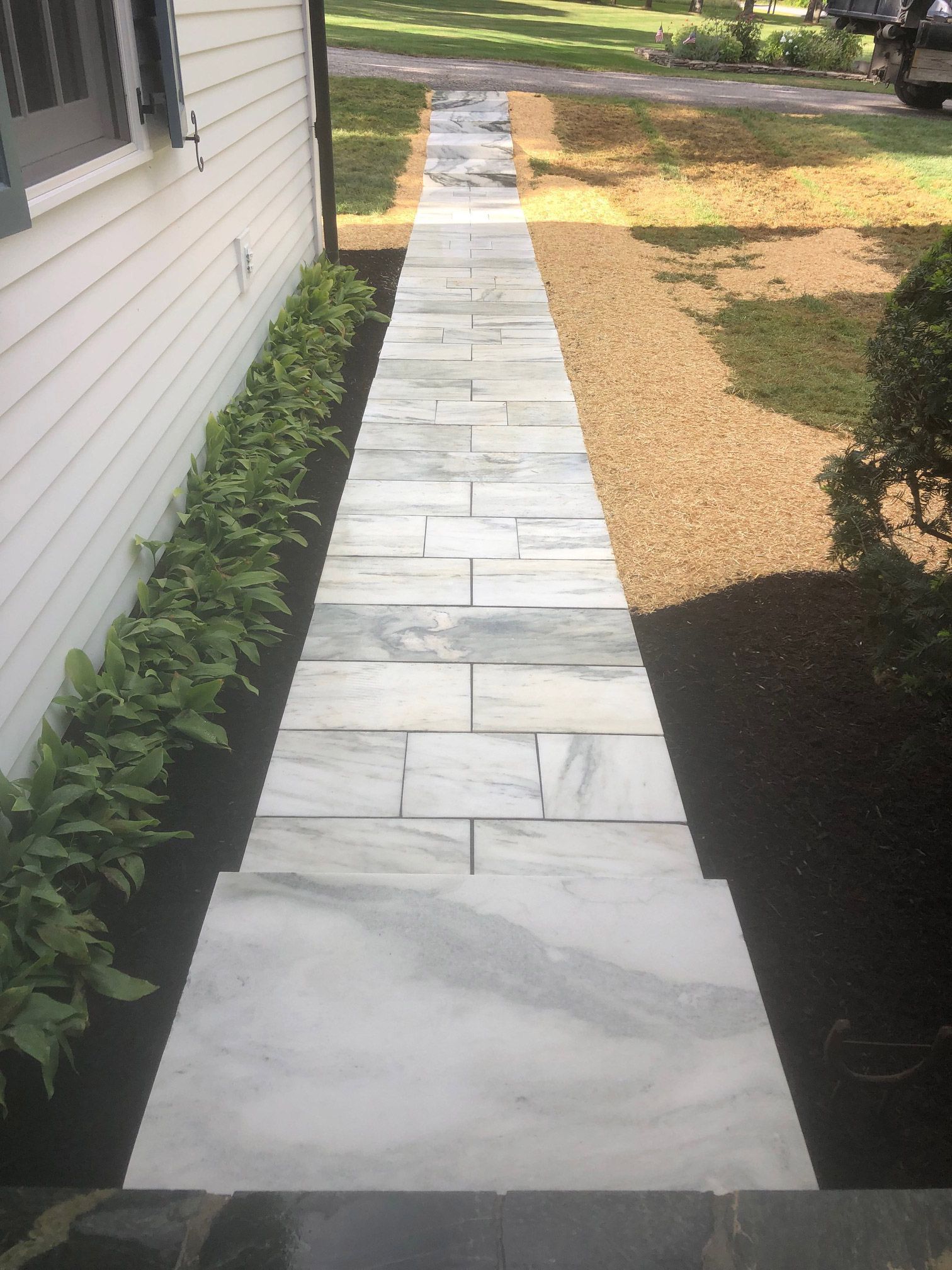 Stone pathway leading from a house, flanked by green plants on one side, and dark mulch and grass.