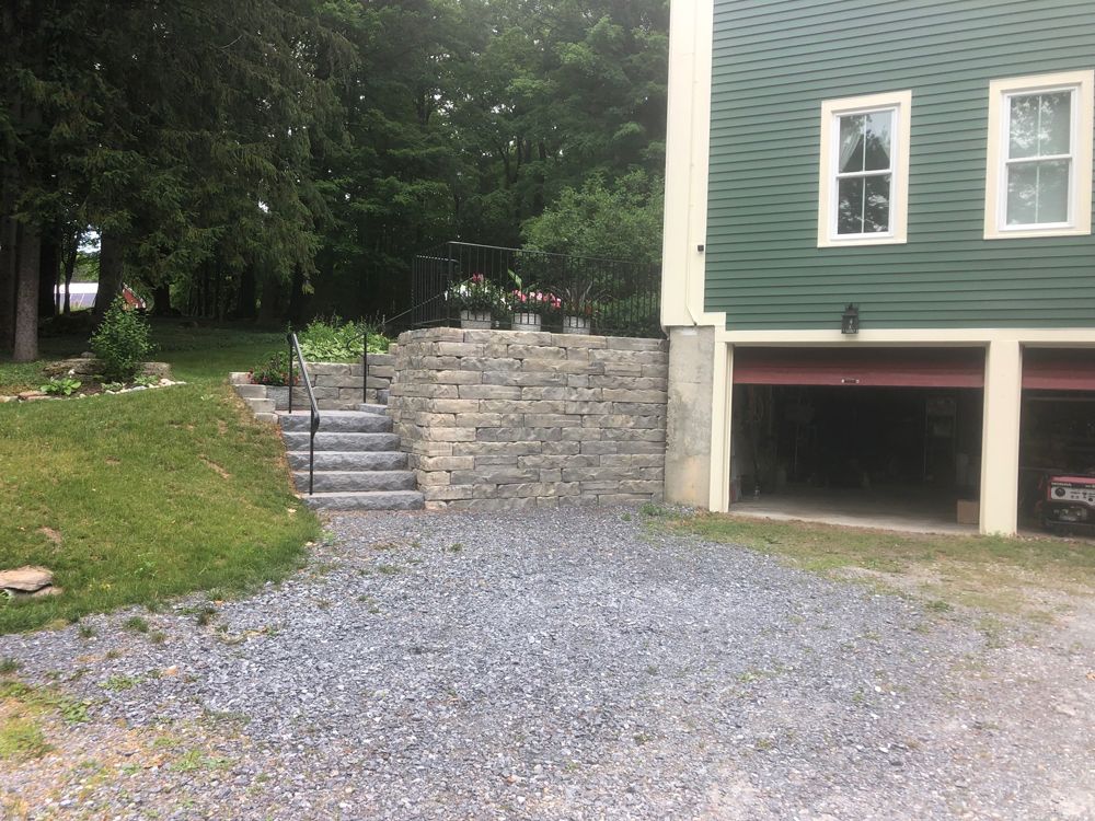 Stone retaining wall with steps, leading to a garden. Green house with open garage doors and gravel driveway.