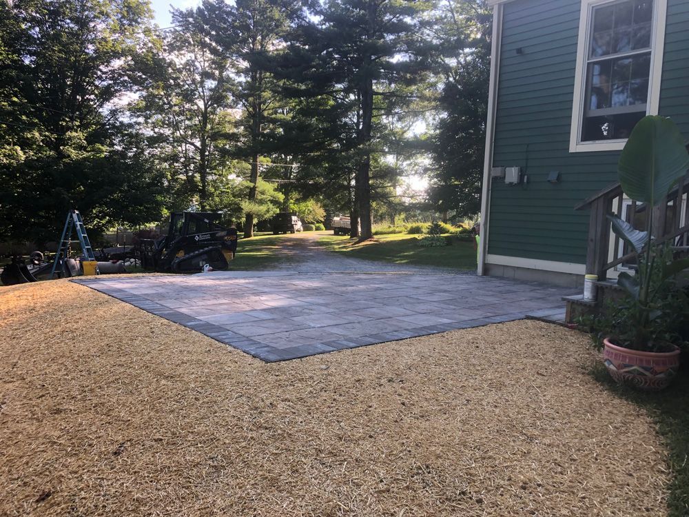 A gravel area and paved patio next to a green house, trees in the background.